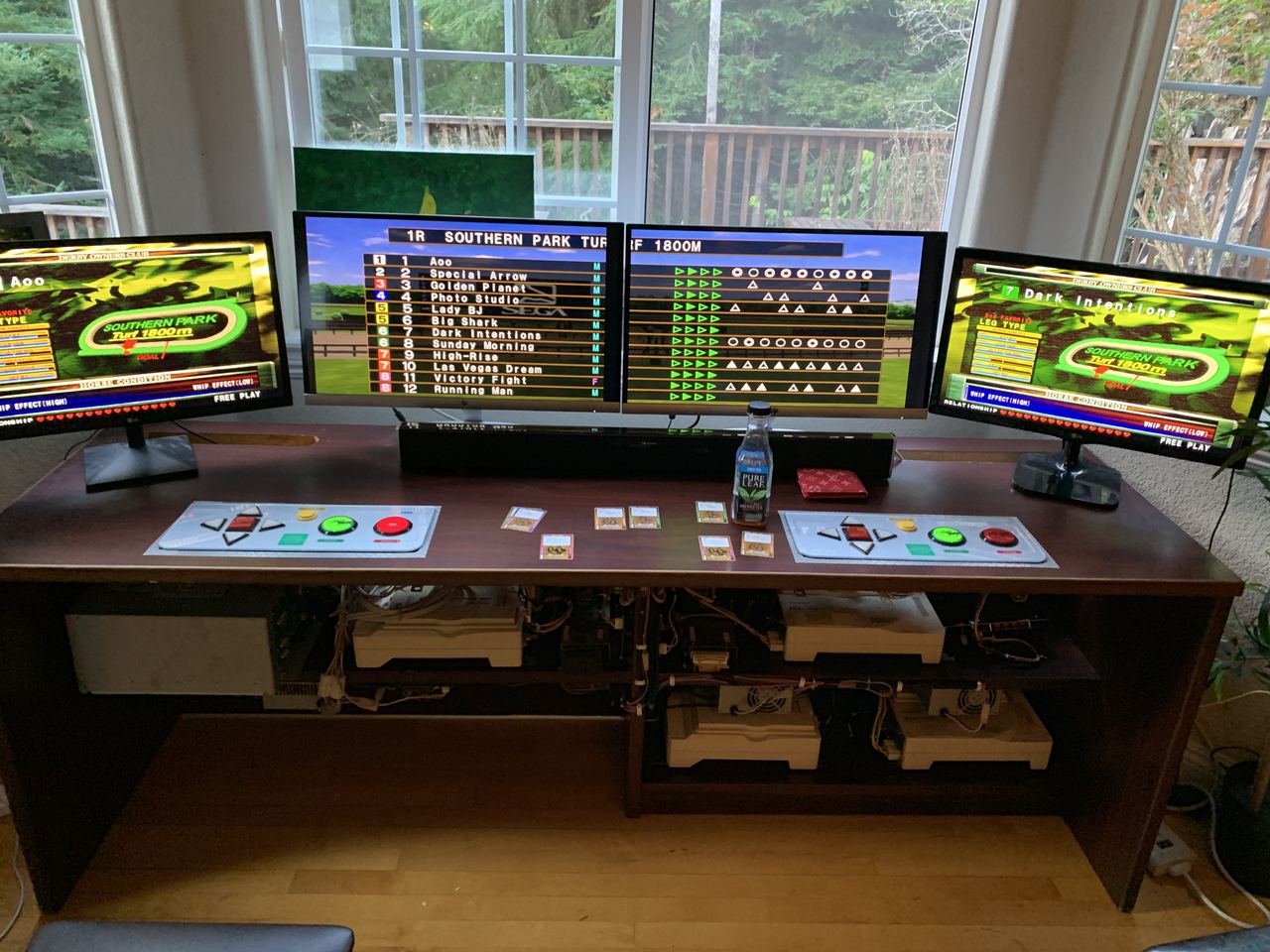 A home rig: two player seats with arcade control panels and card readers, the two-screen race board between them, horse cards on the desk, arcade boards racked below.