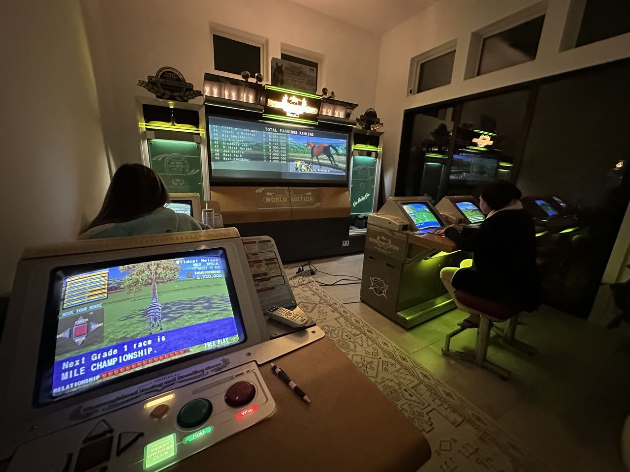 A lit row of Derby Owners Club seats with people playing, the main race board glowing at the end.