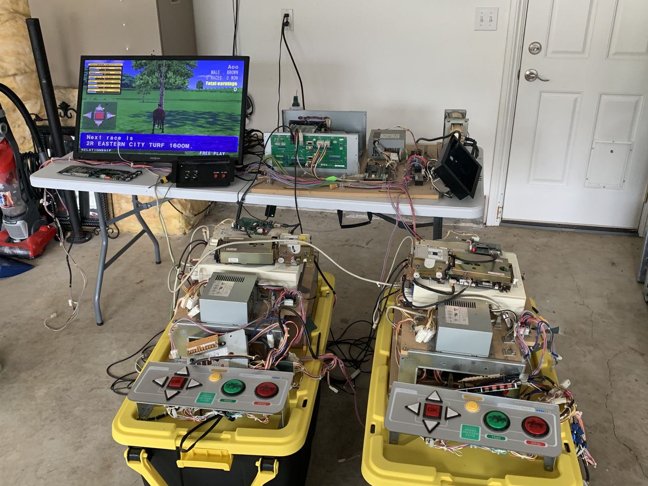 Arcade boards on folding tables in a garage, wired to monitors for testing.
